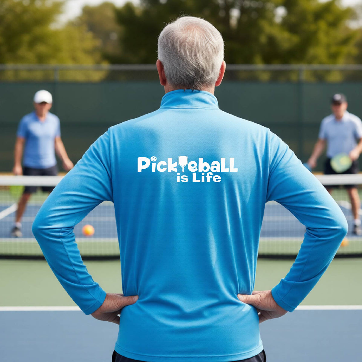 A man viewed from behind on a pickleball court, wearing a bright blue long-sleeve shirt with the white text 'Pickleball is Life' printed across the back.