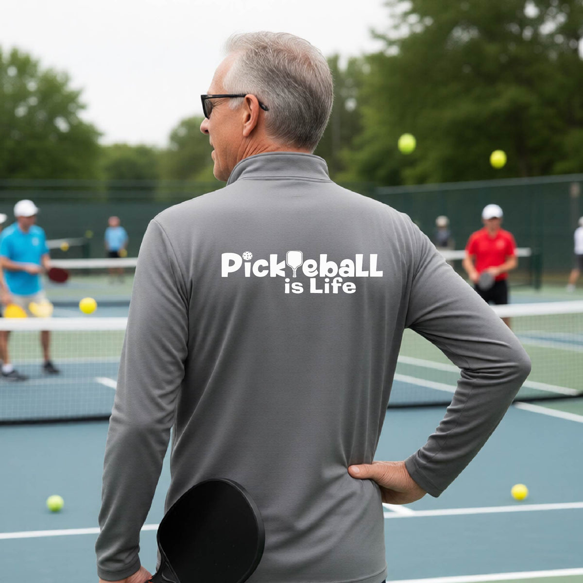 A man viewed from behind on a pickleball court, wearing a gray long-sleeve shirt with the white text 'Pickleball is Life' printed across the back.