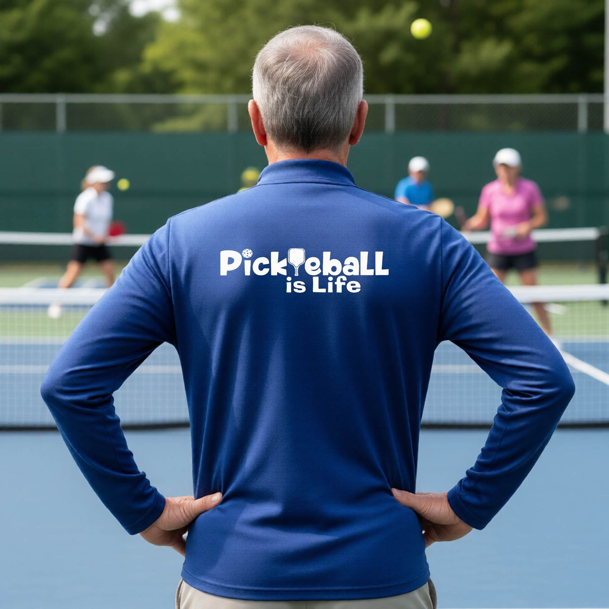 A man viewed from behind on a pickleball court, wearing a royal blue long-sleeve shirt with the white text 'Pickleball is Life' printed across the back.