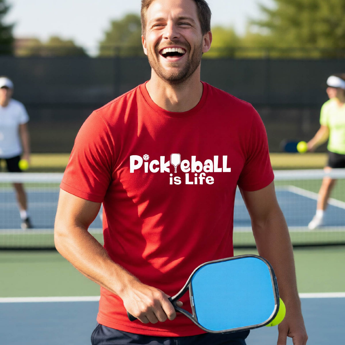A man on a pickleball court wearing a red t-shirt with the text 'Pickleball is Life' in white, with a pickleball paddle in place of the first L.