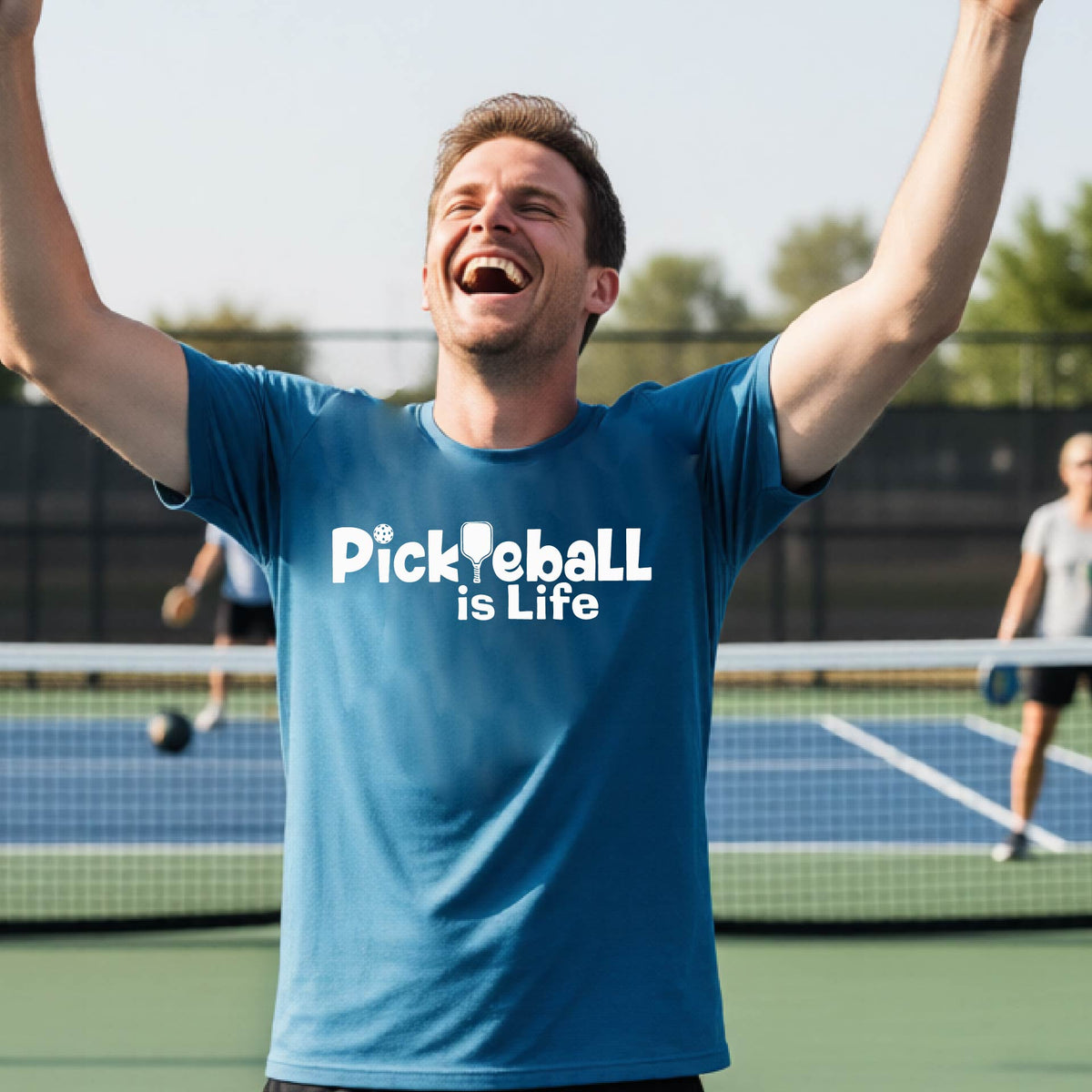 A man on a pickleball court wearing a teal t-shirt with the text 'Pickleball is Life' in white, with a pickleball paddle in place of the first L.