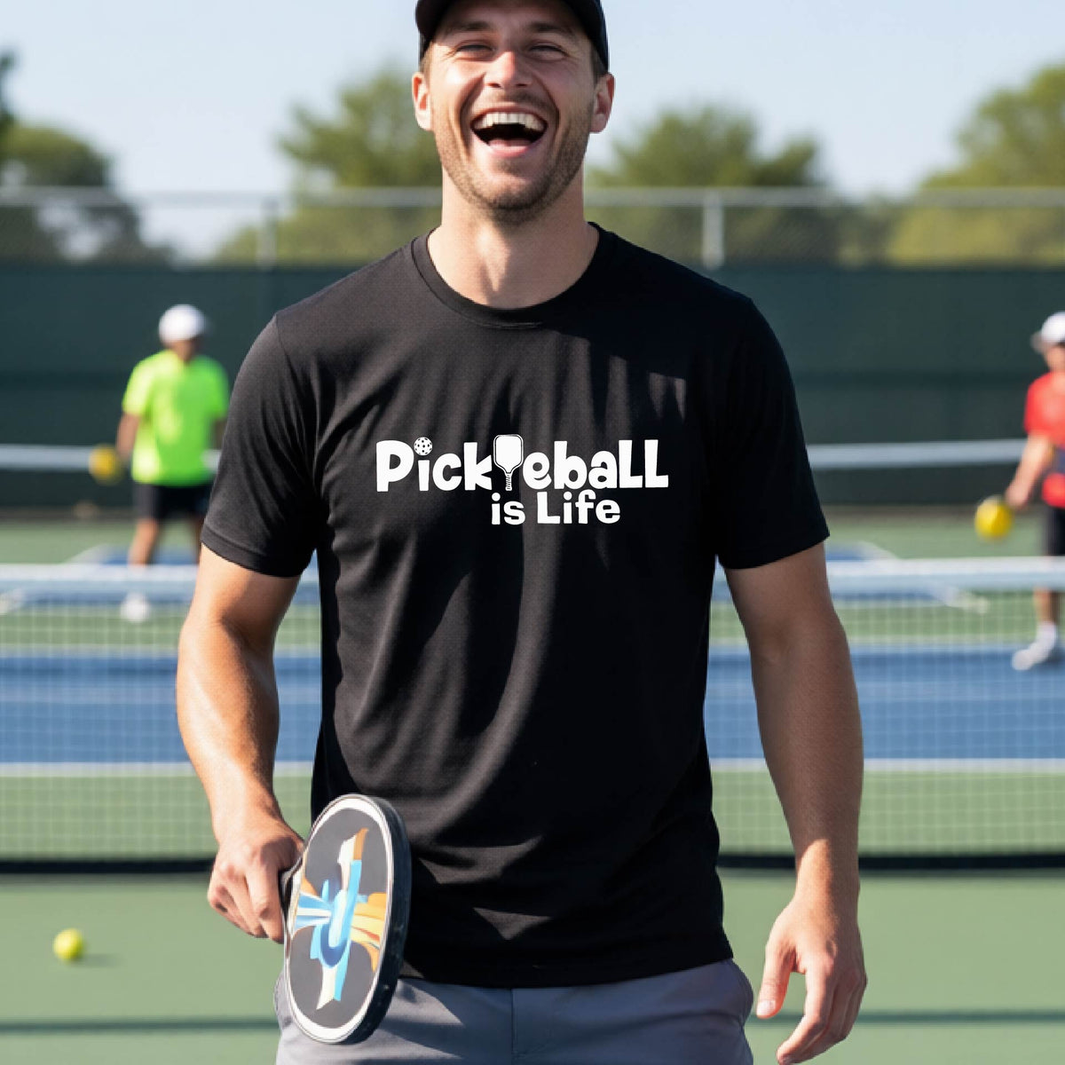A man on a pickleball court wearing a black t-shirt with the text 'Pickleball is Life' in white, with a pickleball paddle in place of the first L.