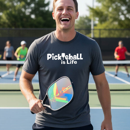 A man on a pickleball court wearing a dark gray t-shirt with the text 'Pickleball is Life' in white, with a pickleball paddle in place of the first L.