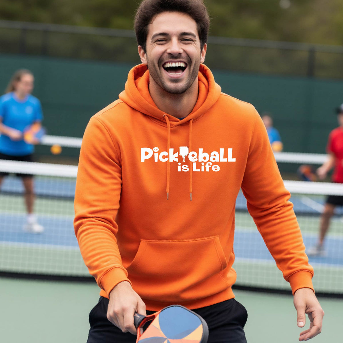 A man wearing an orange hoodie with the text 'Pickleball is Life' in white, standing on a pickleball court .