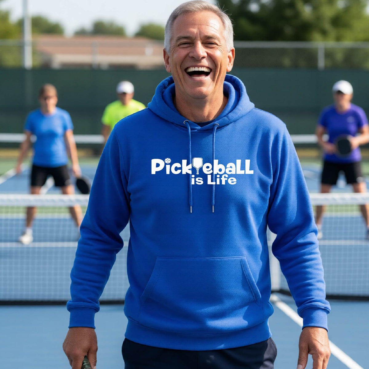 A man wearing a royal blue hoodie with the text 'Pickleball is Life' in white, standing on a pickleball court .