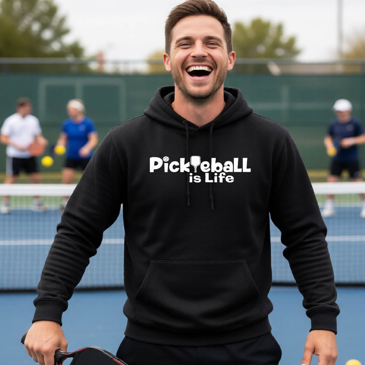 A man wearing a black hoodie with the text 'Pickleball is Life' in white, standing on a pickleball court .