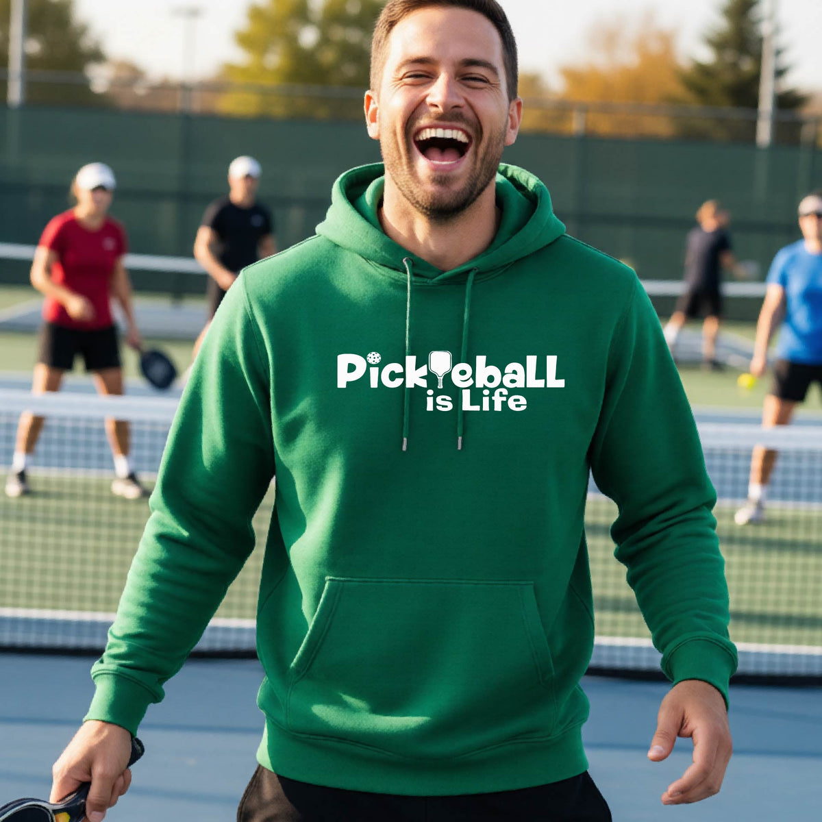 A man wearing a green hoodie with the text 'Pickleball is Life' in white, standing on a pickleball court .