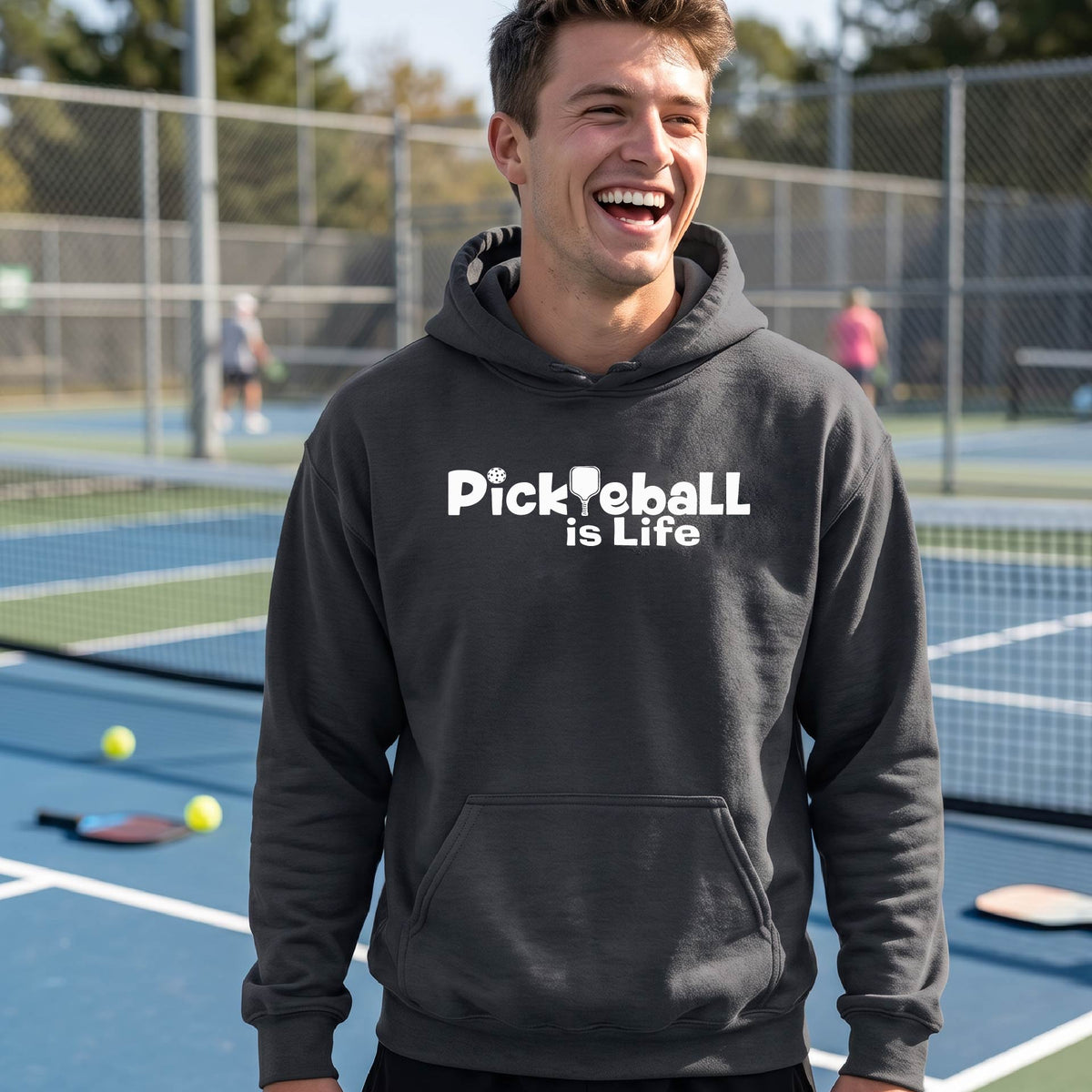 A man wearing a dark gray hoodie with the text 'Pickleball is Life' in white, standing on a pickleball court .