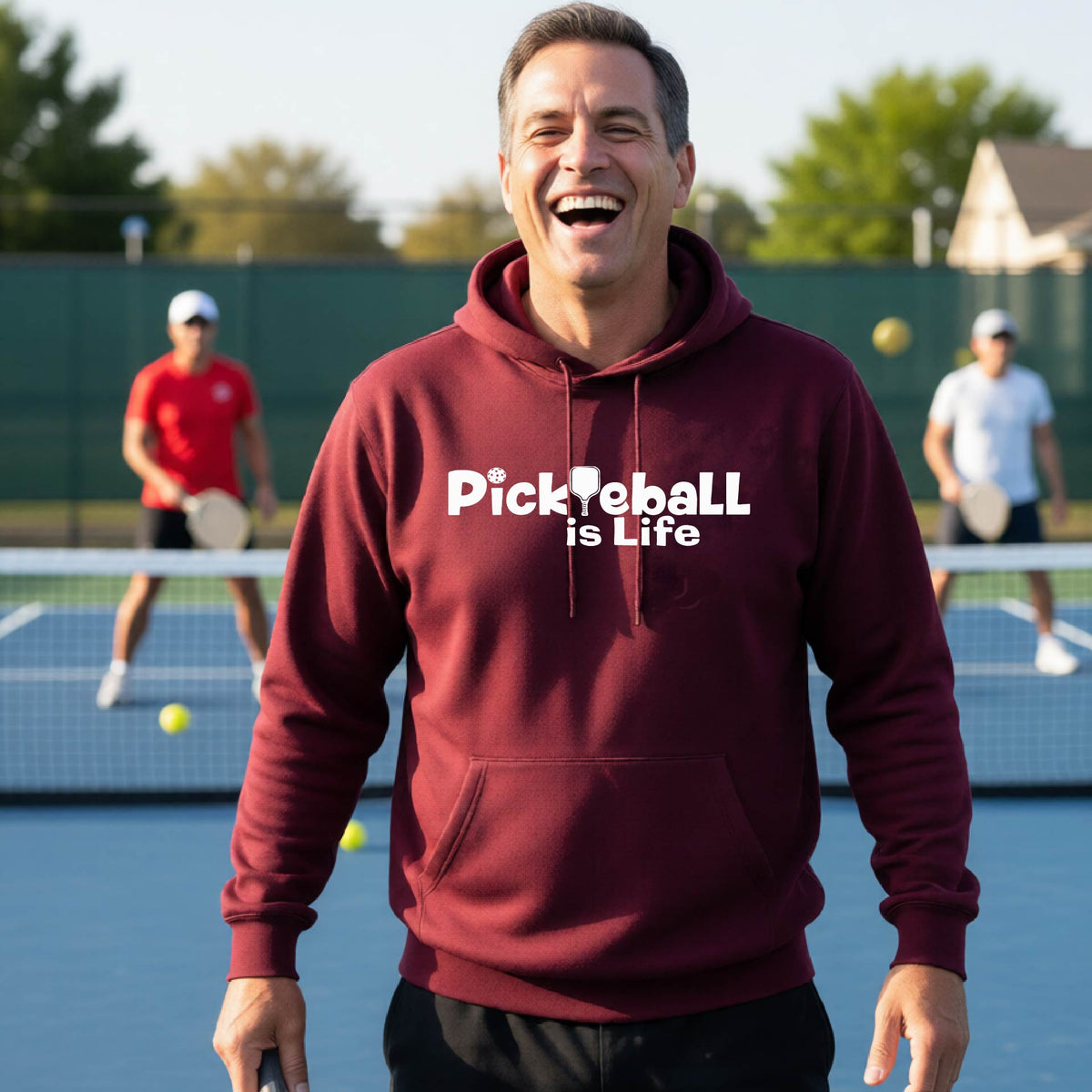 A man wearing a maroon hoodie with the text 'Pickleball is Life' in white, standing on a pickleball court .
