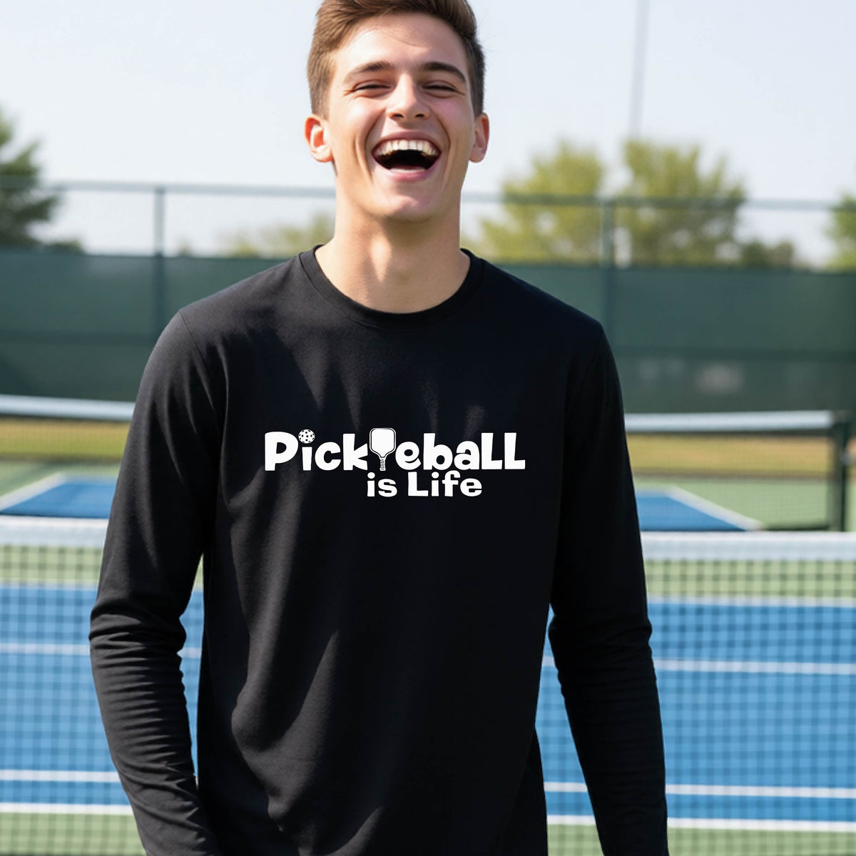 A smiling young person wearing a black long-sleeve t-shirt with the white text 'Pickieball is Life' printed on the chest, with the L replaced by a pickleball paddle.