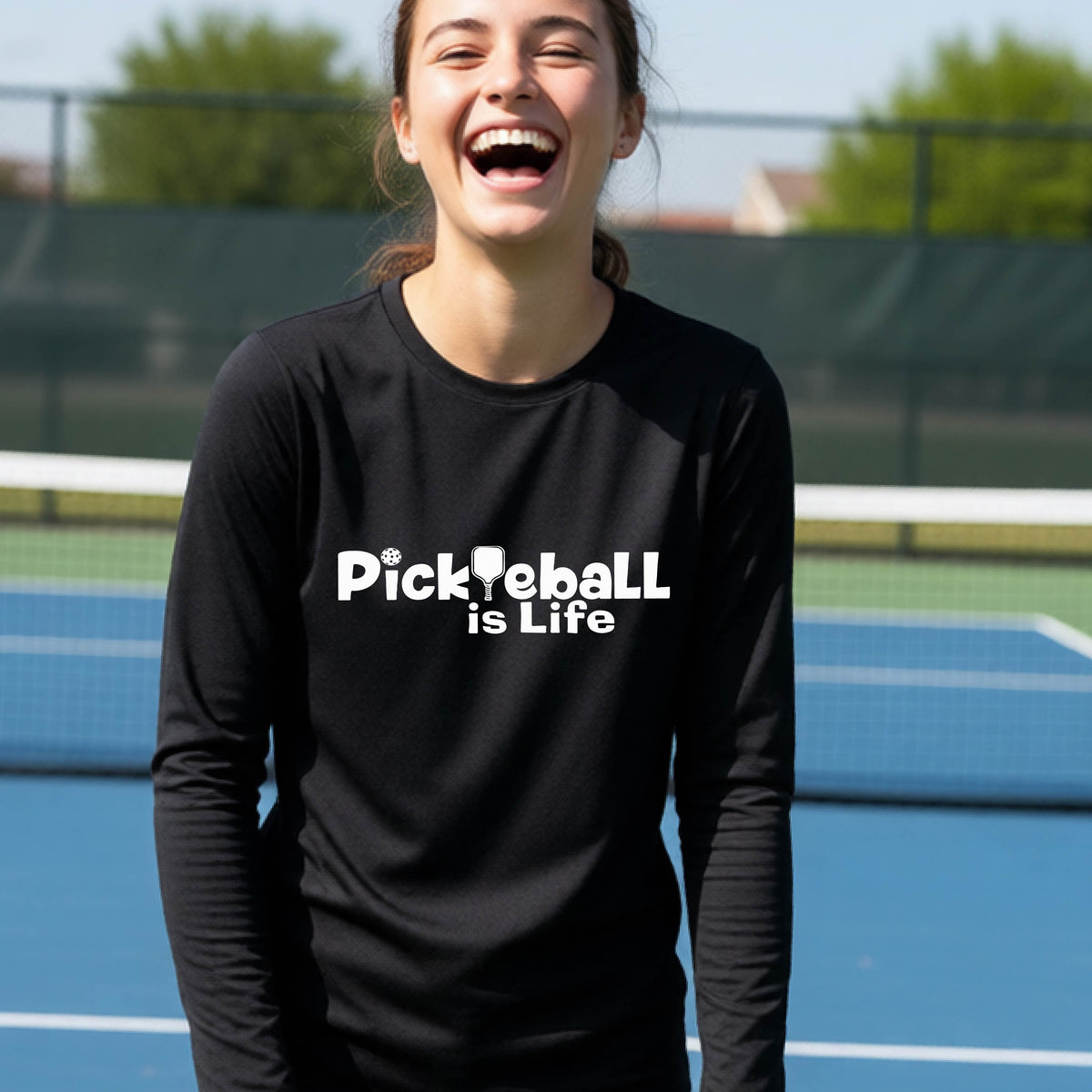 A smiling young person wearing a black long-sleeve t-shirt with the white text 'Pickieball is Life' printed on the chest, with the L replaced by a pickleball paddle.