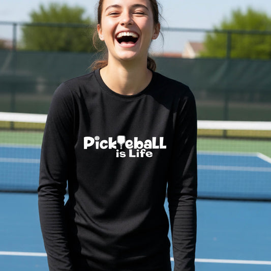 A smiling young person wearing a black long-sleeve t-shirt with the white text 'Pickieball is Life' printed on the chest, with the L replaced by a pickleball paddle.
