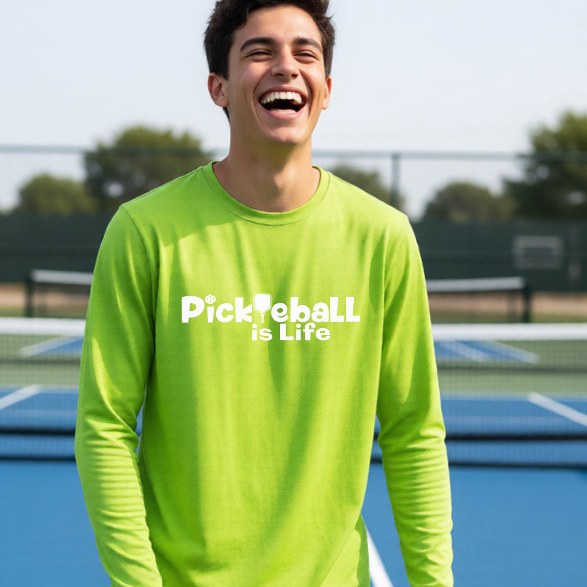 A smiling young person wearing a lime green long-sleeve t-shirt with the white text 'Pickieball is Life' printed on the chest, with the L replaced by a pickleball paddle.