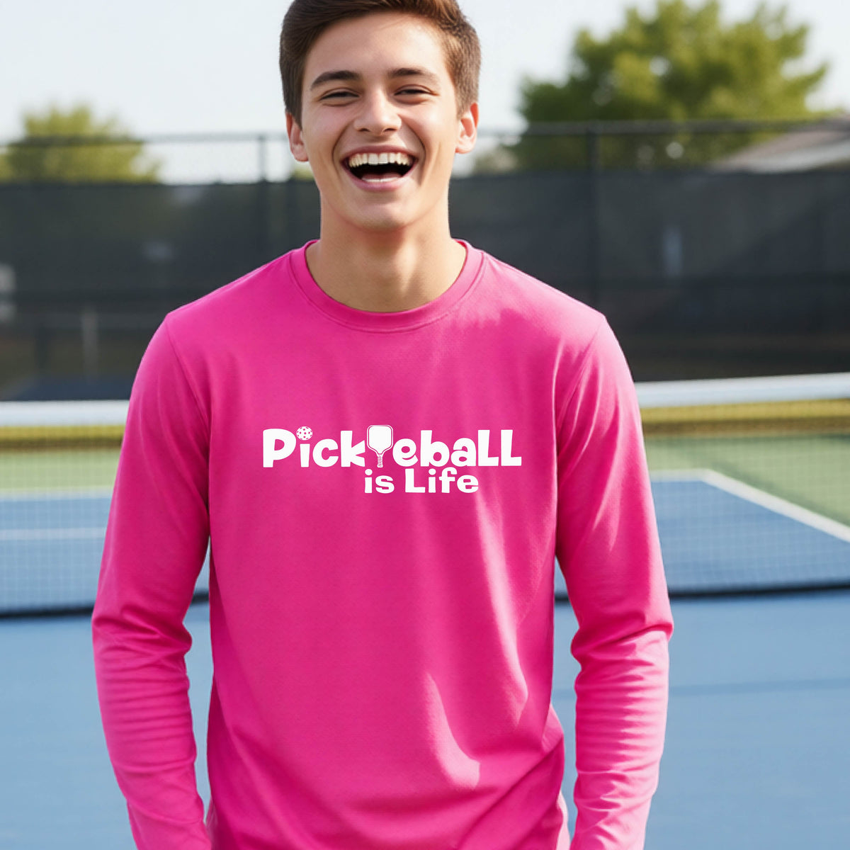 A smiling young person wearing a bright pink long-sleeve t-shirt with the white text 'Pickieball is Life' printed on the chest, with the L replaced by a pickleball paddle.