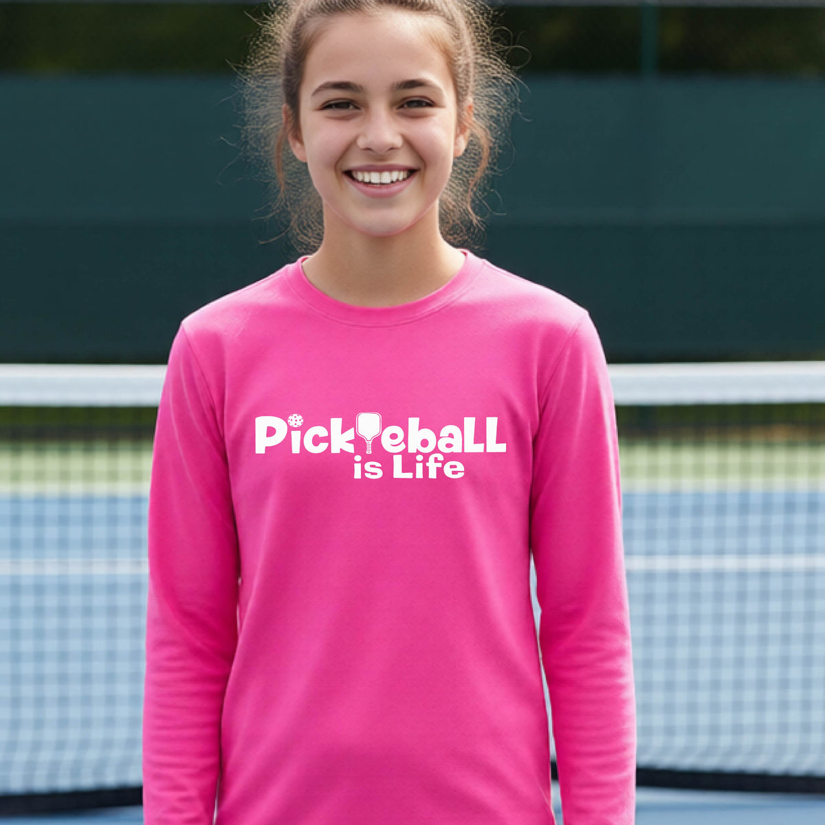 A smiling young person wearing a bright pink long-sleeve t-shirt with the white text 'Pickieball is Life' printed on the chest, with the L replaced by a pickleball paddle.