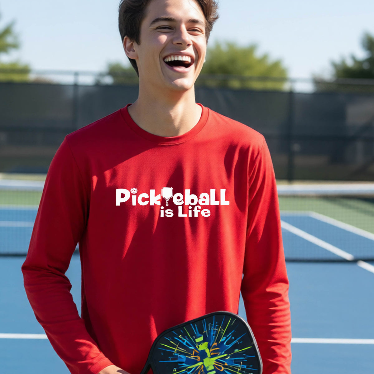 A smiling young person wearing a bright red long-sleeve t-shirt with the white text 'Pickieball is Life' printed on the chest, with the L replaced by a pickleball paddle.