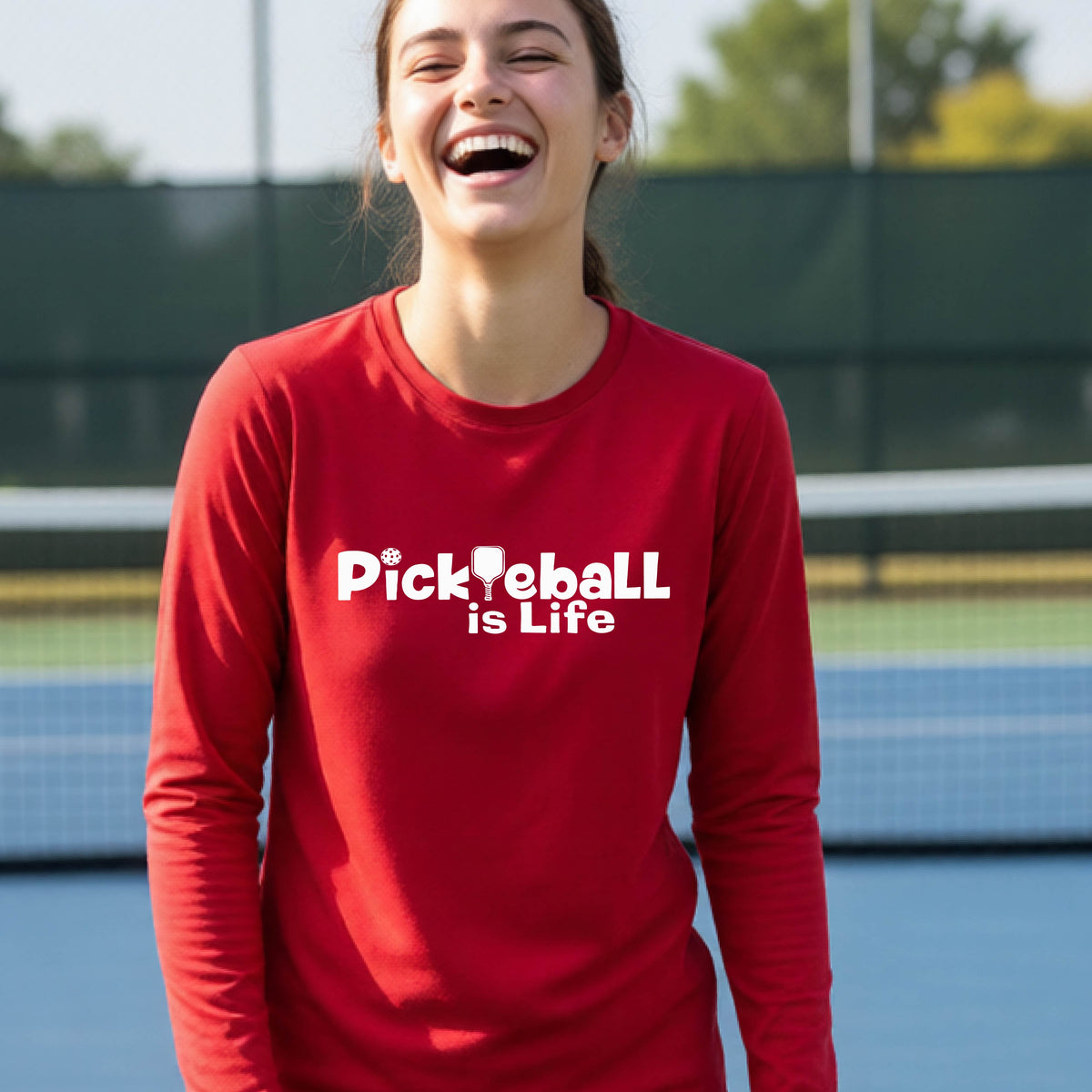 A smiling young person wearing a bright red long-sleeve t-shirt with the white text 'Pickieball is Life' printed on the chest, with the L replaced by a pickleball paddle.