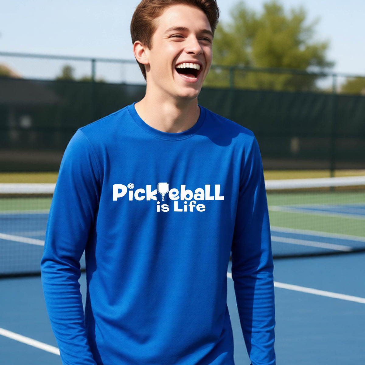 A smiling young person wearing a bright royal  long-sleeve t-shirt with the white text 'Pickieball is Life' printed on the chest, with the L replaced by a pickleball paddle.
