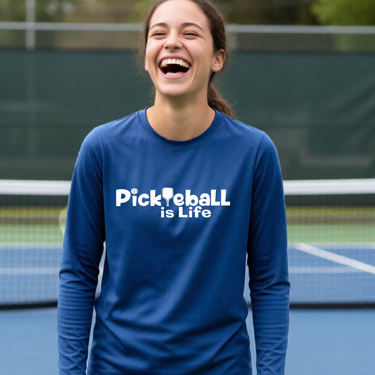 A smiling young person wearing a royal blue long-sleeve t-shirt with the white text 'Pickieball is Life' printed on the chest, with the L replaced by a pickleball paddle.