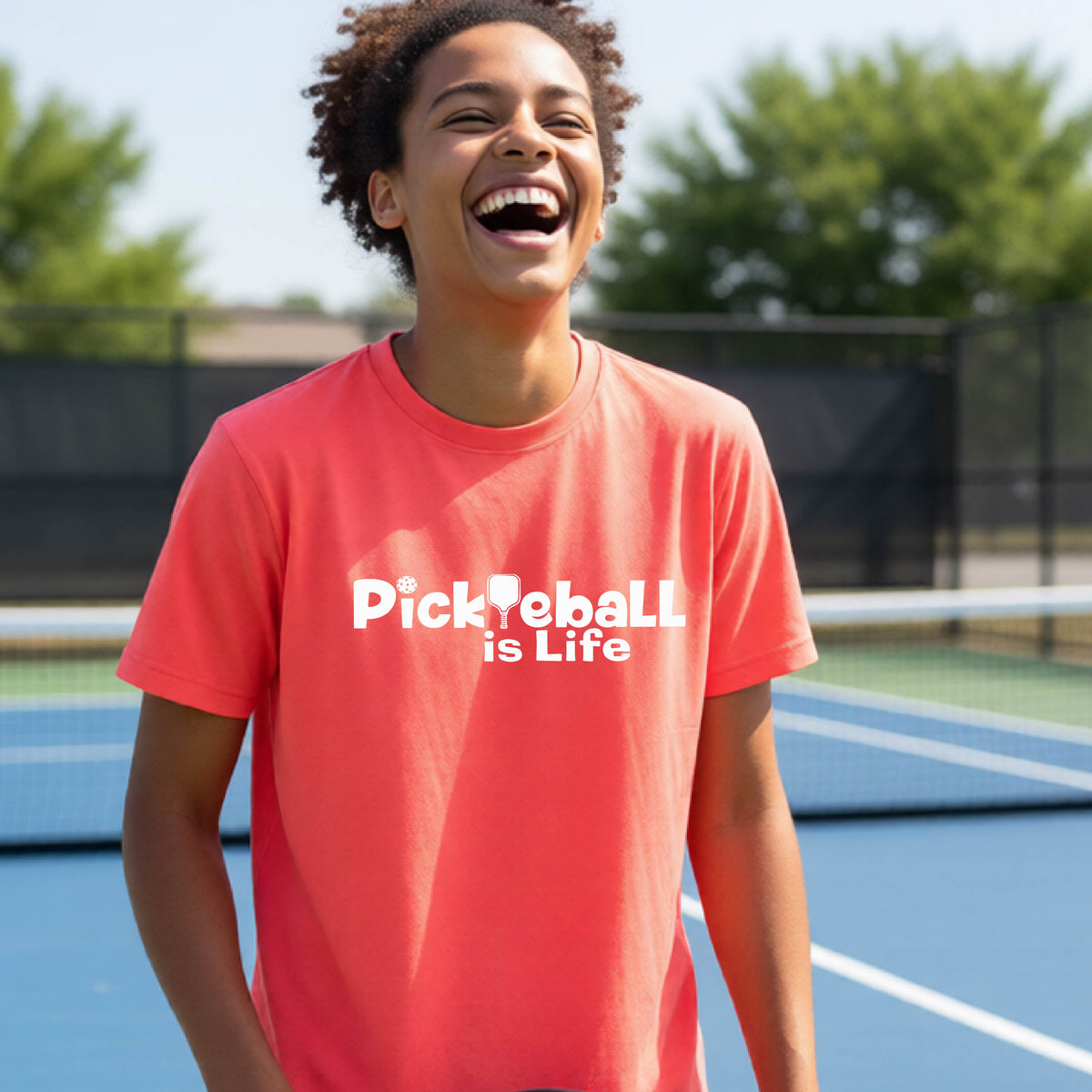 A young teenager laughing on a pickleball court, wearing a coral pink t-shirt with the white text 'Pickleball is Life' with replaced by a pickleball paddle the first L across the chest.