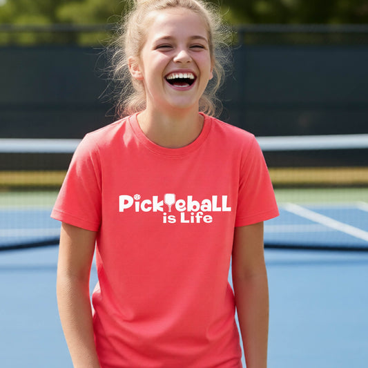 A young teenager laughing on a blue pickleball court, wearing a coral pink t-shirt with the white text 'Pickleball is Life' with replaced by a pickleball paddle the first L across the chest.