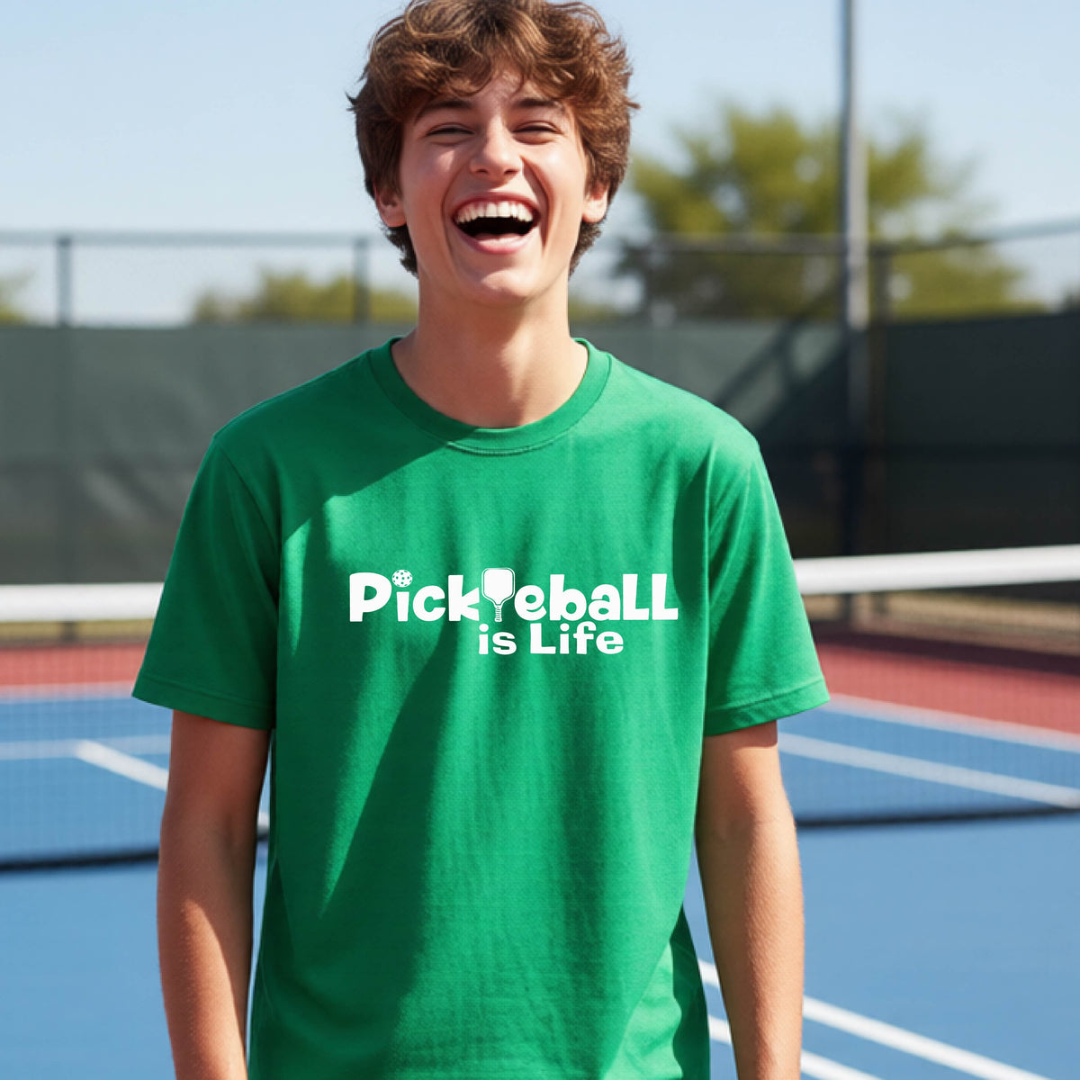 A young teenager laughing on a pickleball court, wearing a green t-shirt with the white text 'Pickleball is Life' with replaced by a pickleball paddle the first L across the chest.