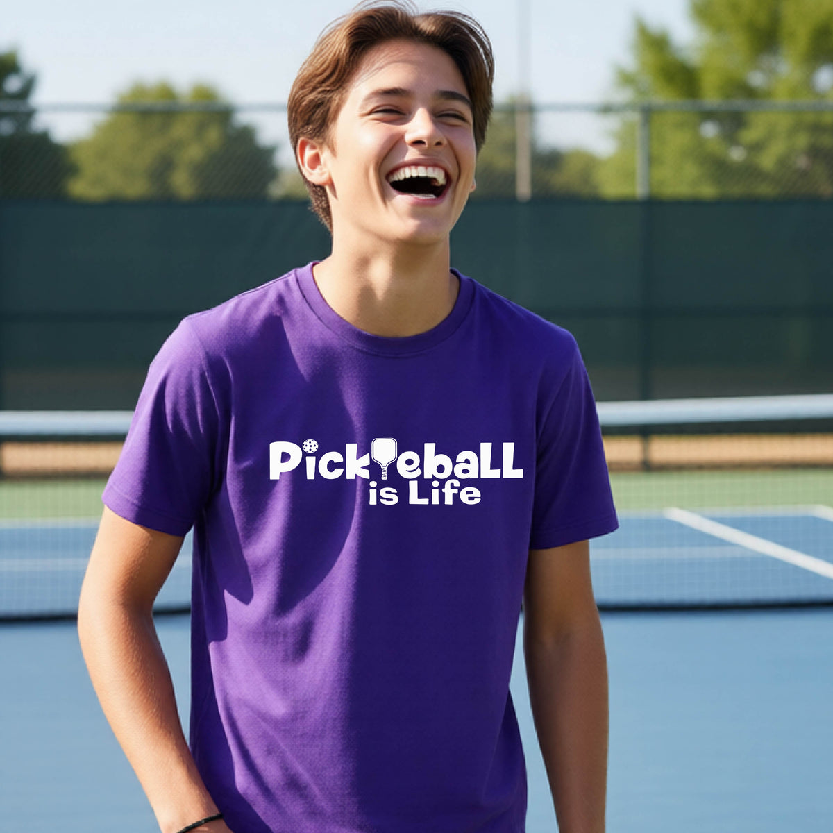 A young teenager laughing on a pickleball court, wearing a purple t-shirt with the white text 'Pickleball is Life' with replaced by a pickleball paddle the first L across the chest.