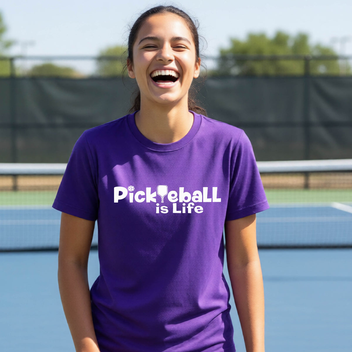 A young teenager laughing on a pickleball court, wearing a purple t-shirt with the white text 'Pickleball is Life' with replaced by a pickleball paddle the first L across the chest.
