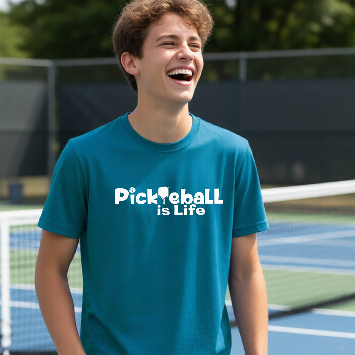 A young teenager laughing on a pickleball court, wearing a teal t-shirt with the white text 'Pickleball is Life' with replaced by a pickleball paddle the first L across the chest.