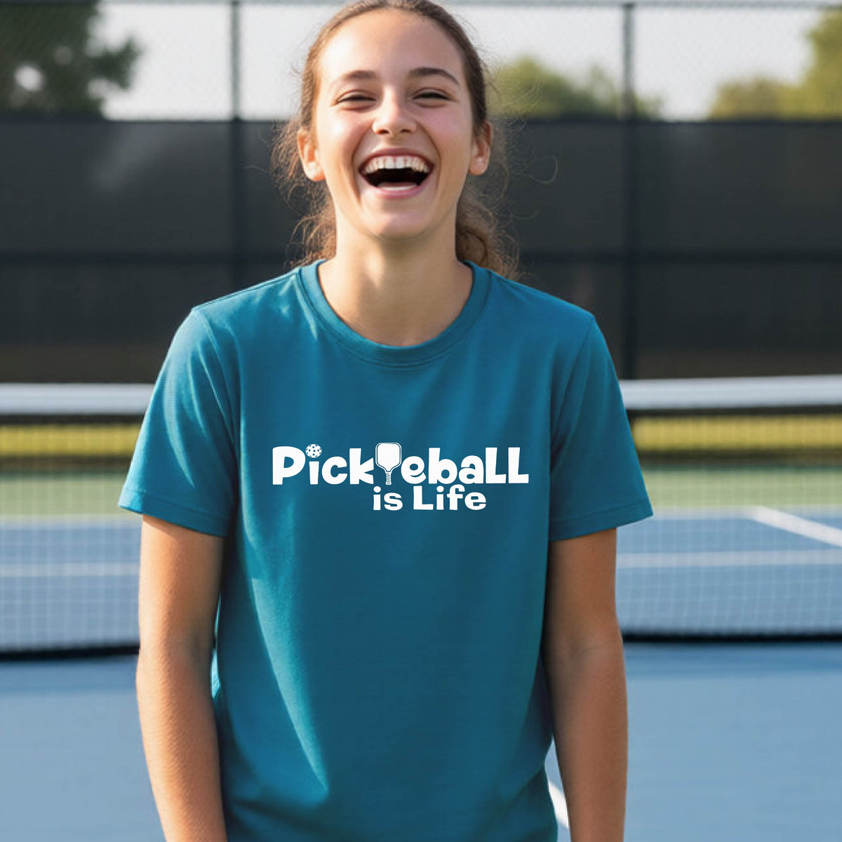 A young teenager laughing on a pickleball court, wearing a teal t-shirt with the white text 'Pickleball is Life' with replaced by a pickleball paddle the first L across the chest.