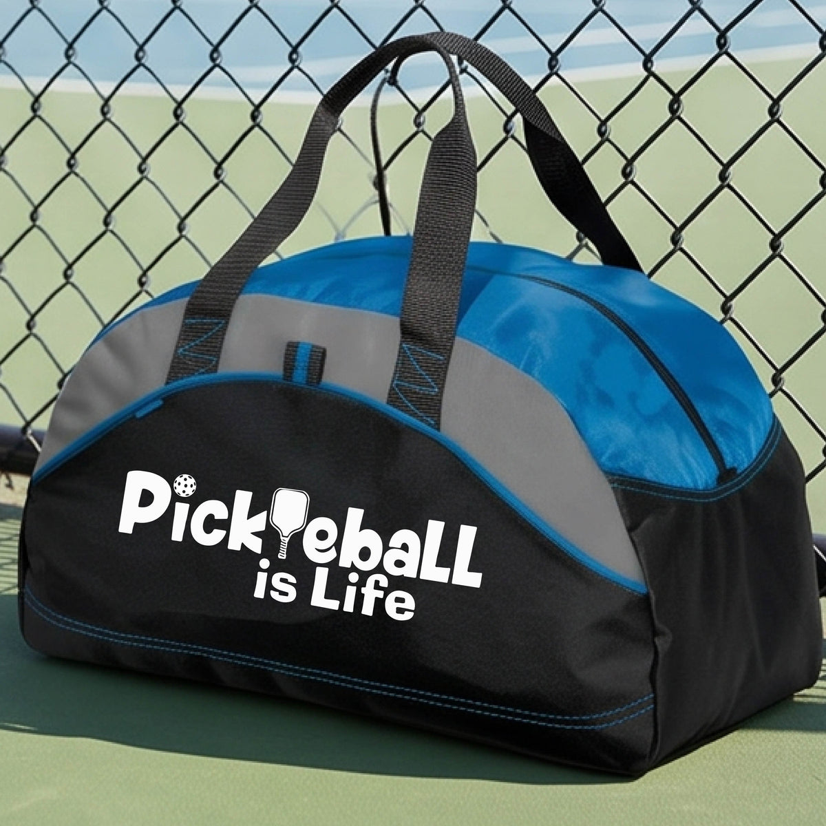 A royal blue, black, and gray duffel bag with the text 'Pickbeall is Life' printed on the front, placed on a tennis court.