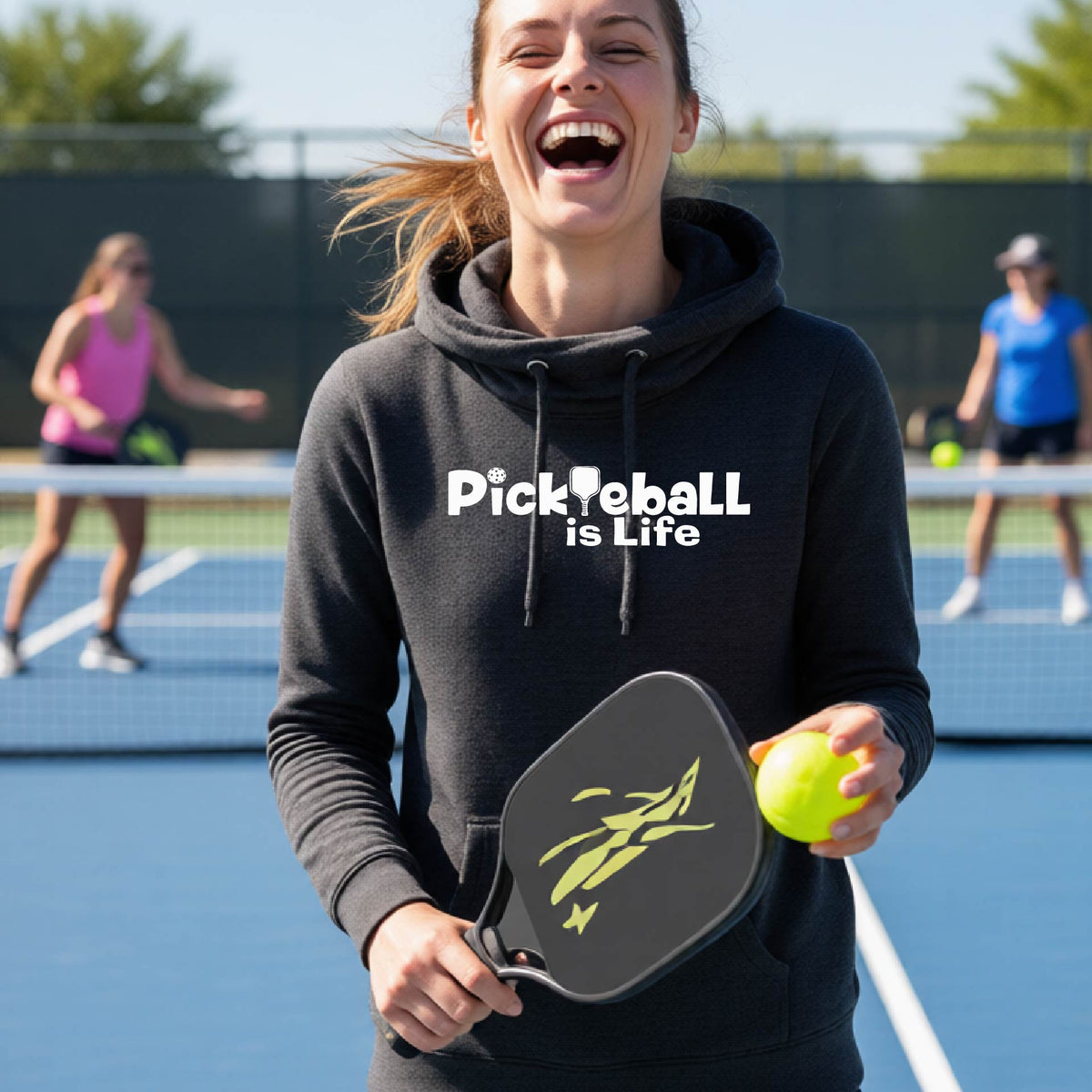 A woman on a pickleball court wearing a black hoodie with the text 'Pickleball is Life'with the L replaced with a pickleball paddle, graphic.