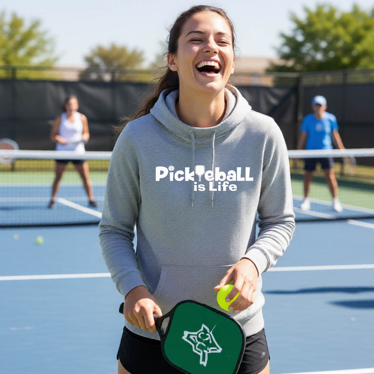 A woman on a pickleball court wearing a gray hoodie with the text 'Pickleball is Life'with the L replaced with a pickleball paddle, graphic.