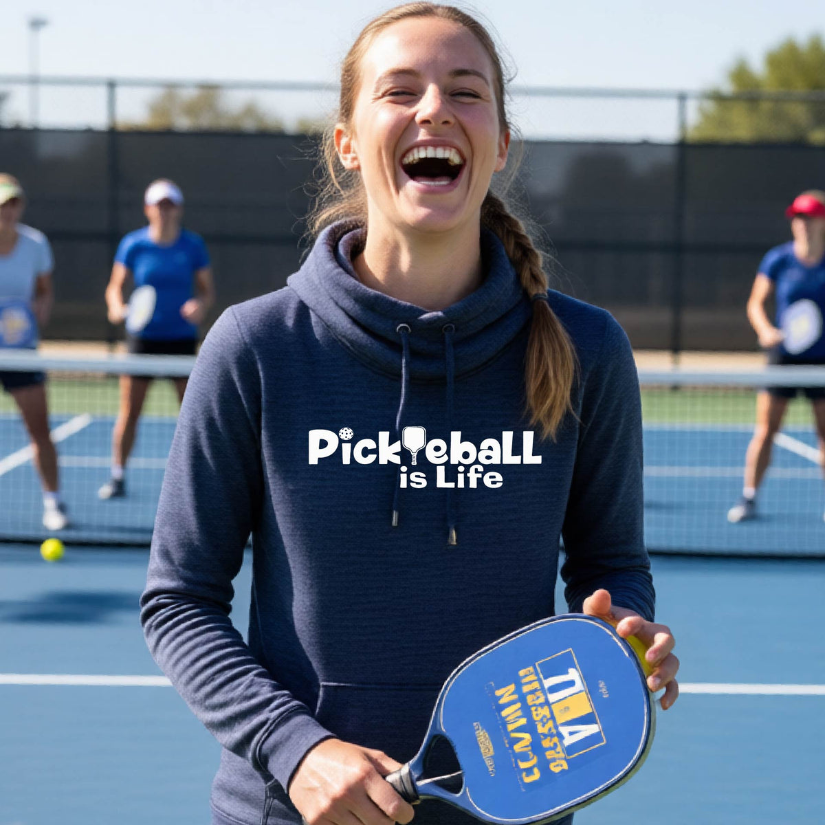 A woman on a pickleball court wearing a navy blue hoodie with the text 'Pickleball is Life'with the L replaced with a pickleball paddle, graphic.
