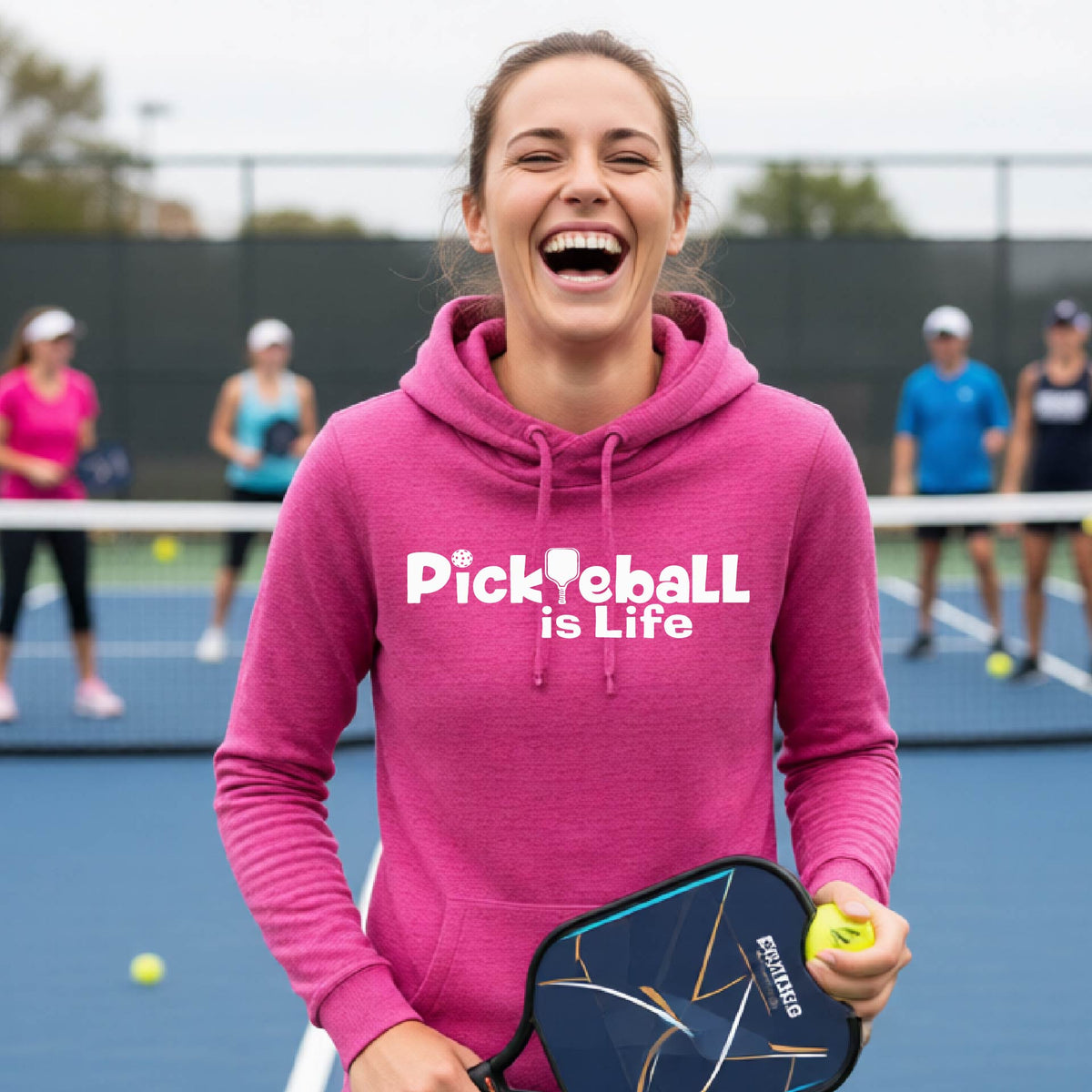 A woman on a pickleball court wearing a pink hoodie with the text 'Pickleball is Life'with the L replaced with a pickleball paddle, graphic.