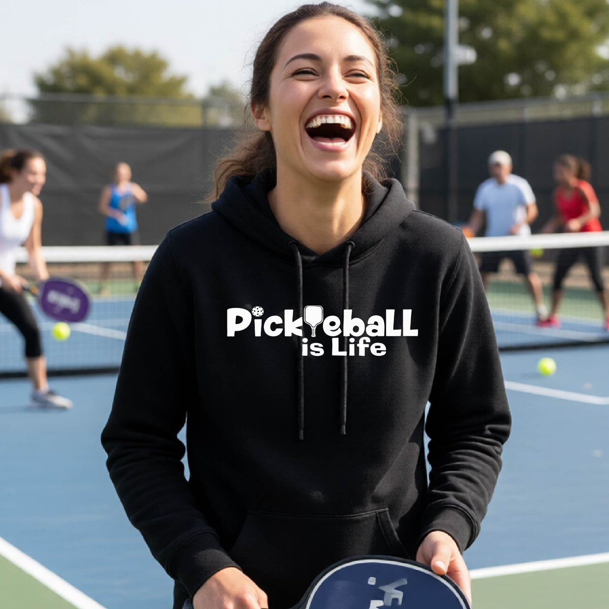 A woman laughing on an outdoor pickleball court wearing a black hoodie with the text 'Pickleball is Life' printed in white across the chest.
