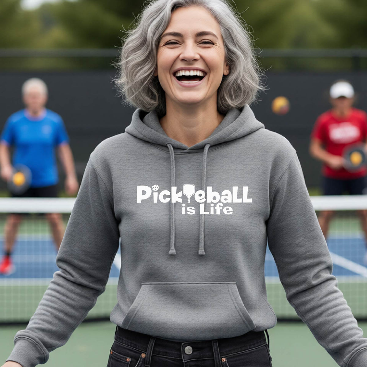 A woman laughing on an outdoor pickleball court wearing a gray hoodie with the text 'Pickleball is Life' printed in white across the chest.