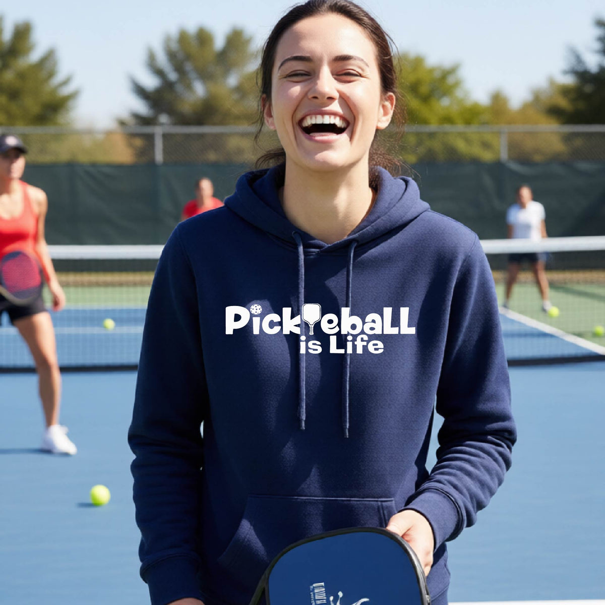 A woman laughing on an outdoor pickleball court wearing a navy blue hoodie with the text 'Pickleball is Life' printed in white across the chest.