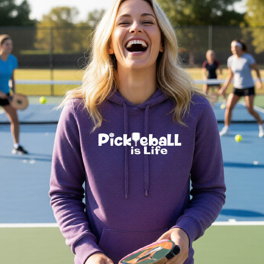 A woman laughing on an outdoor pickleball court wearing a purple hoodie with the text 'Pickleball is Life' printed in white across the chest.