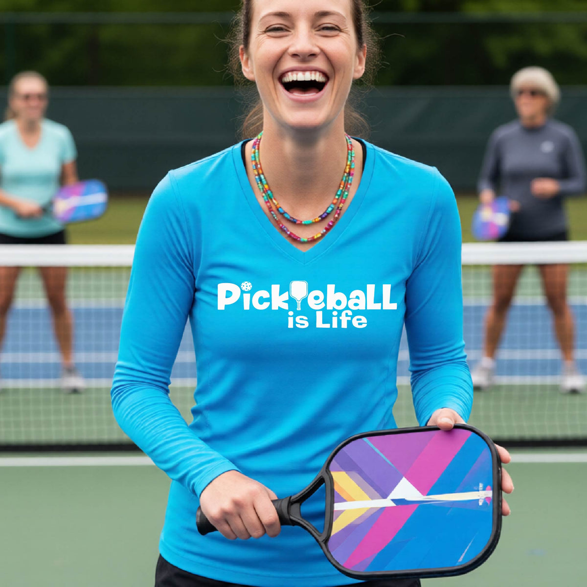 A woman wearing a bright blue long-sleeve v-neck t-shirt with the slogan 'Pickleball is Life' printed in white across the back, with a pickleball paddle replacing the L.