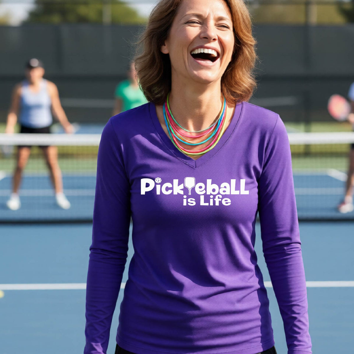 A woman wearing a purple long-sleeve v-neck t-shirt with the slogan 'Pickleball is Life' printed in white across the back, with a pickleball paddle replacing the L.