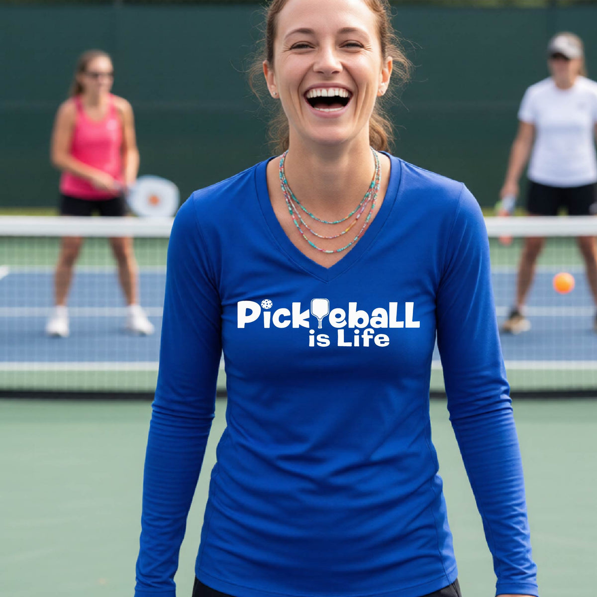A woman wearing a royal blue long-sleeve v-neck t-shirt with the slogan 'Pickleball is Life' printed in white across the back, with a pickleball paddle replacing the L.