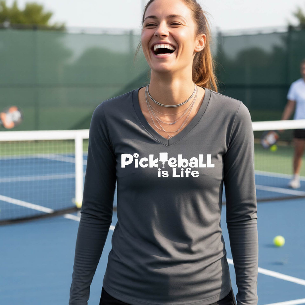 A woman wearing a dark gray long-sleeve v-neck t-shirt with the slogan 'Pickleball is Life' printed in white across the back, with a pickleball paddle replacing the L.