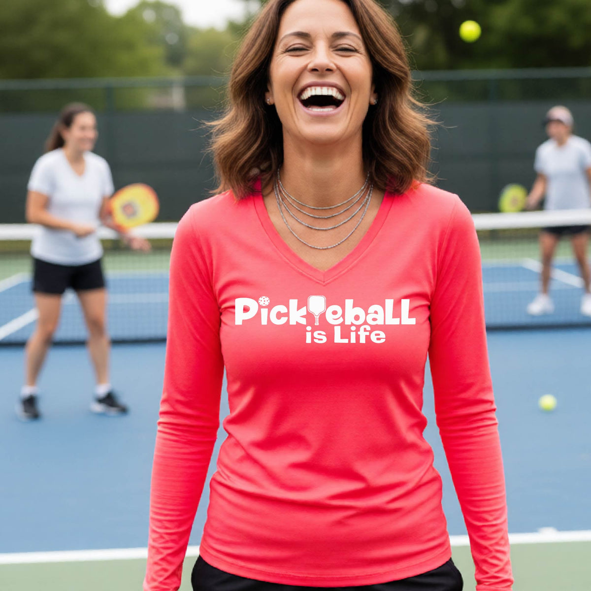 A woman wearing a coral pink long-sleeve v-neck t-shirt with the slogan 'Pickleball is Life' printed in white across the back, with a pickleball paddle replacing the L.
