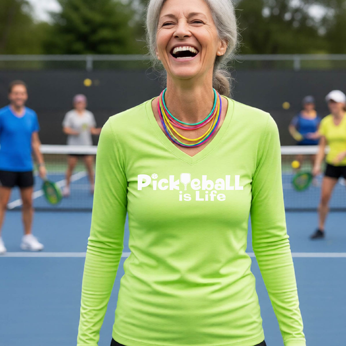 A woman wearing a lime green long-sleeve v-neck t-shirt with the slogan 'Pickleball is Life' printed in white across the back, with a pickleball paddle replacing the L.