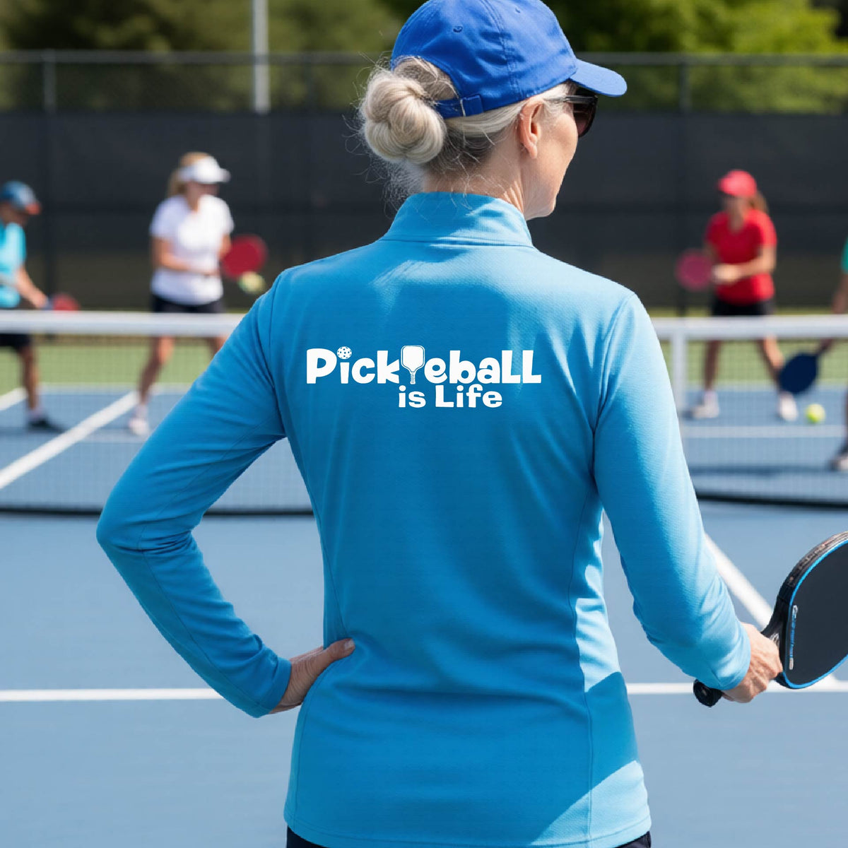 A person on a pickleball court seen from behind wearing a bright blue long-sleeve quarter-zip shirt with the slogan 'Pickleball is Life' printed in white across the back, with a pickleball paddle replacing the L. 