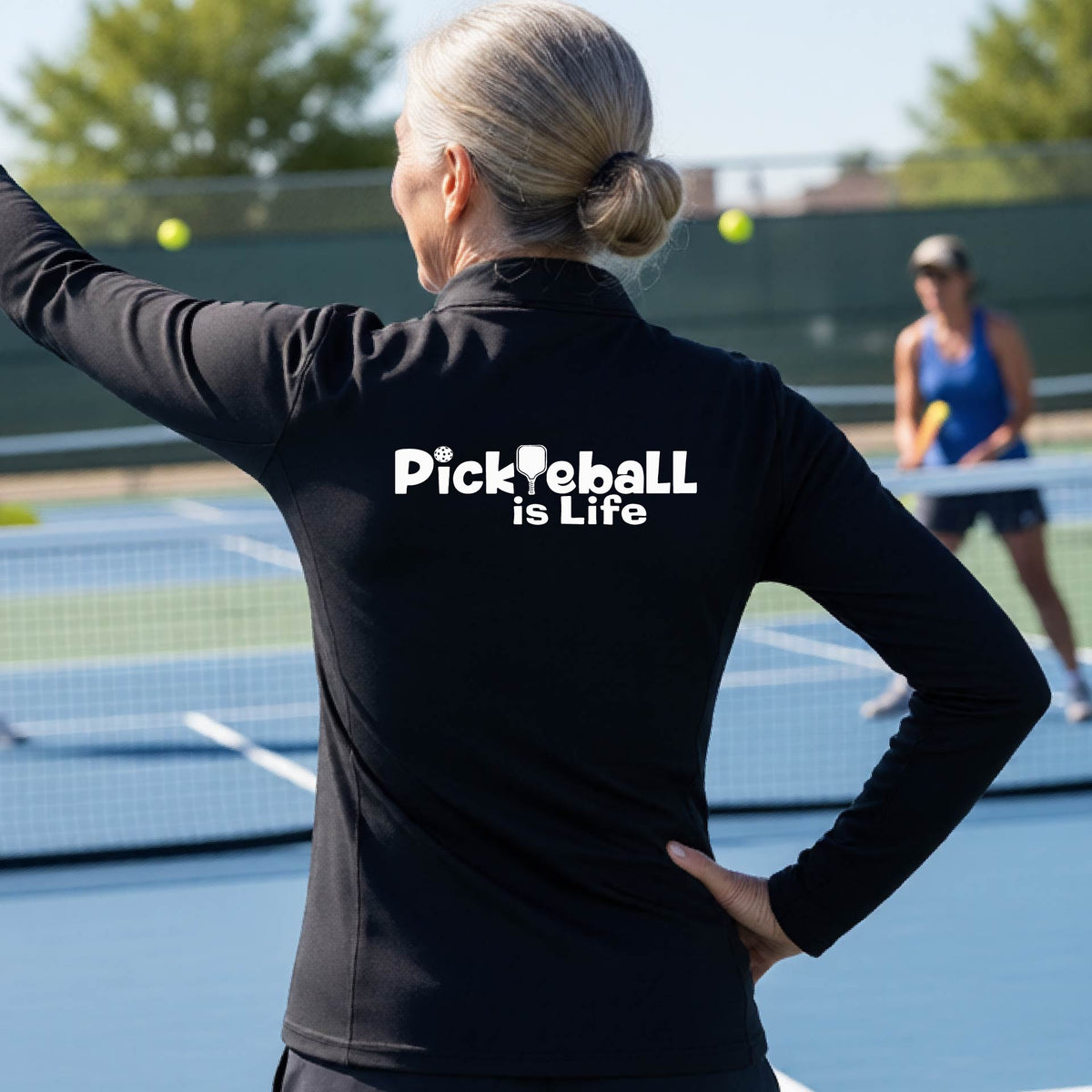 A person on a pickleball court seen from behind wearing a black long-sleeve quarter-zip shirt with the slogan 'Pickleball is Life' printed in white across the back, with a pickleball paddle replacing the L. 