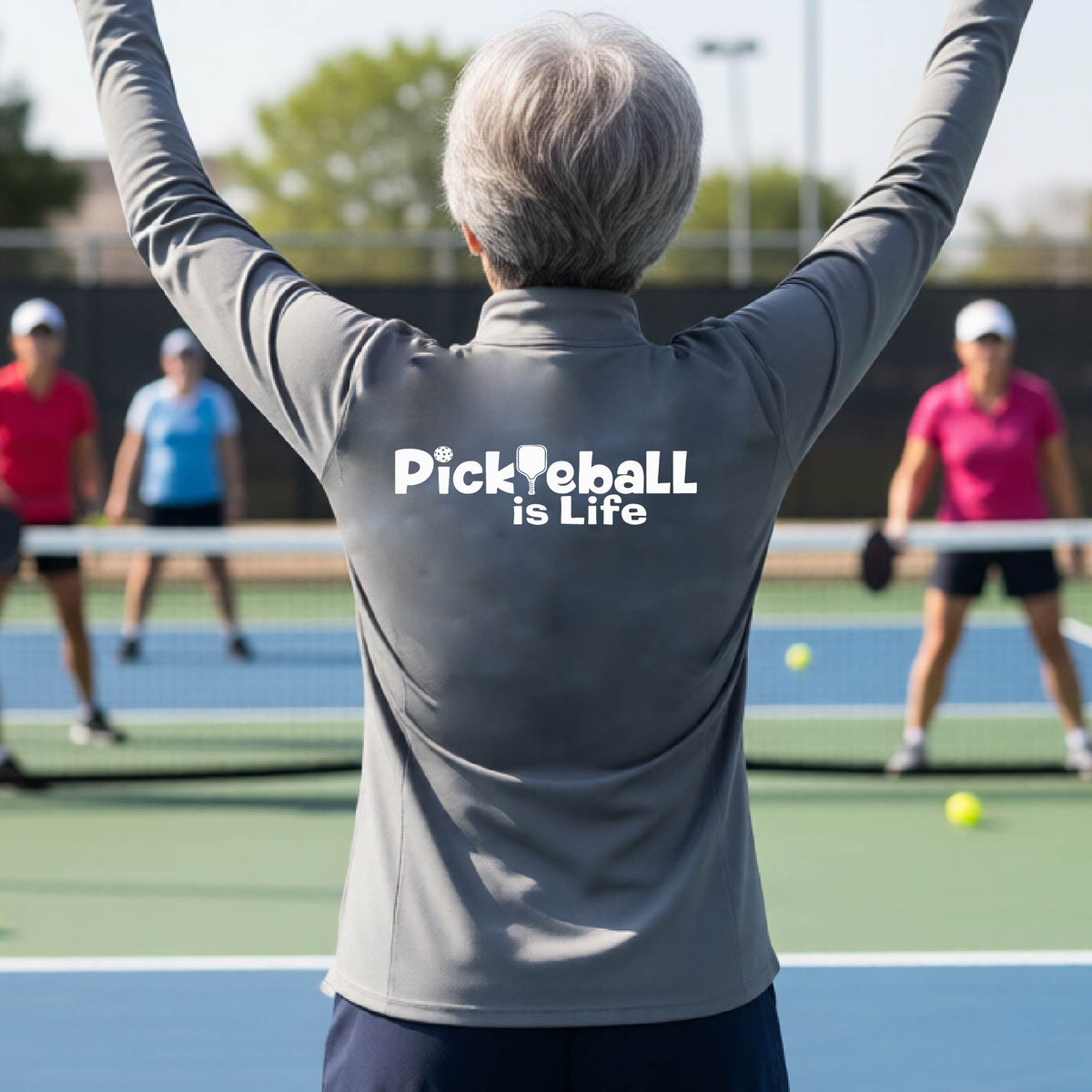 A person on a pickleball court seen from behind wearing a gray long-sleeve quarter-zip shirt with the slogan 'Pickleball is Life' printed in white across the back, with a pickleball paddle replacing the L. 