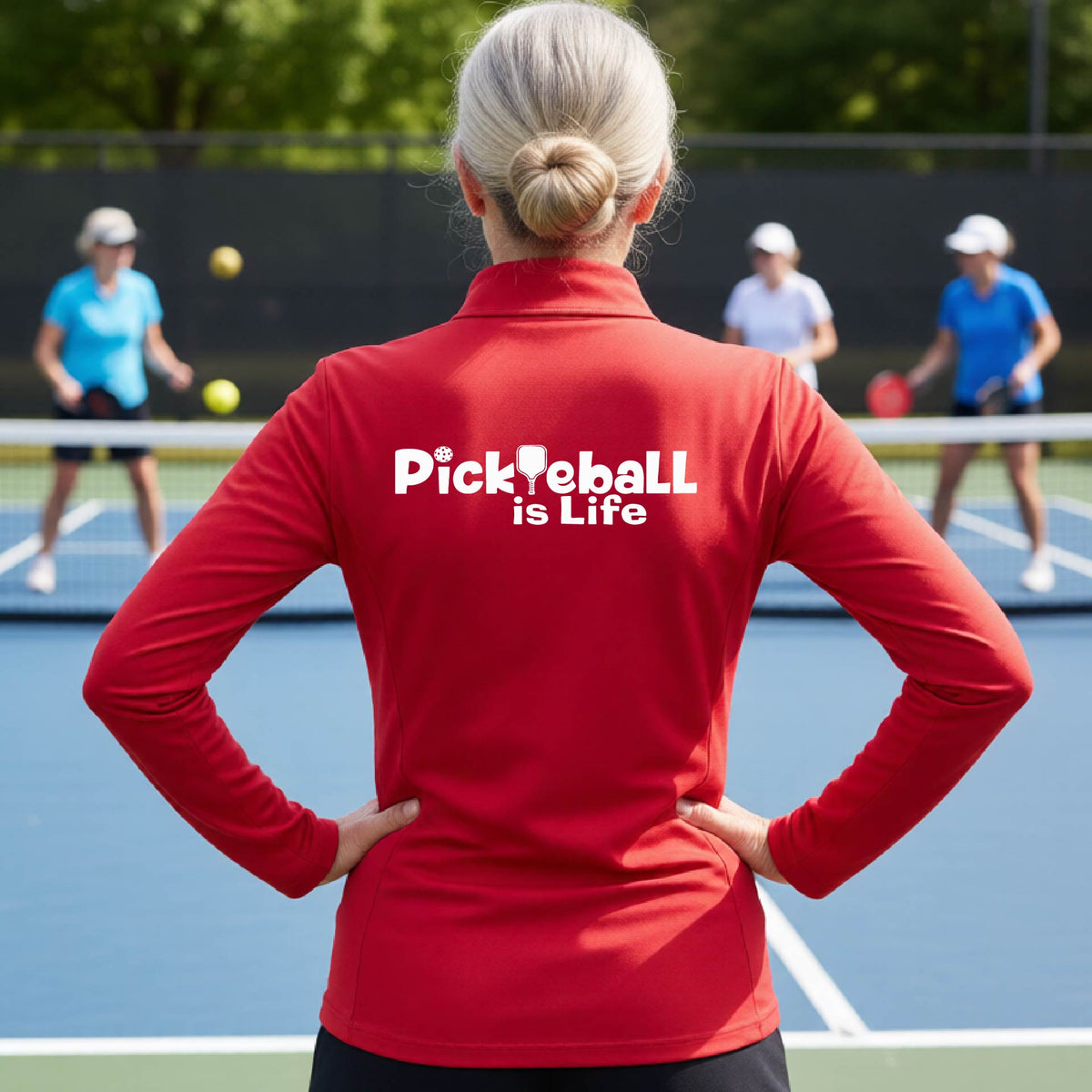 A person on a pickleball court seen from behind wearing a red long-sleeve quarter-zip shirt with the slogan 'Pickleball is Life' printed in white across the back, with a pickleball paddle replacing the L. 