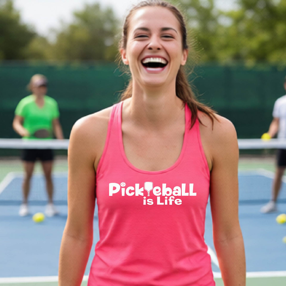 A woman wearing a coral pink racerback tank top with the slogan 'Pickleball is Life' printed in white across the back, with a pickleball paddle replacing the L. 